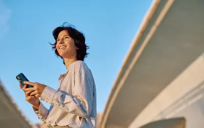 Femme souriante, avec un smartphone en main, sous un ciel bleu.