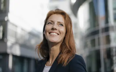 Femme souriante en blazer devant un bâtiment, regarde vers l'avenir.