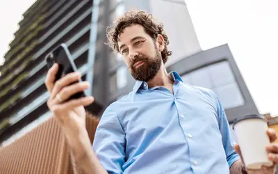 Un homme consulte son téléphone et tient un café devant un bâtiment.