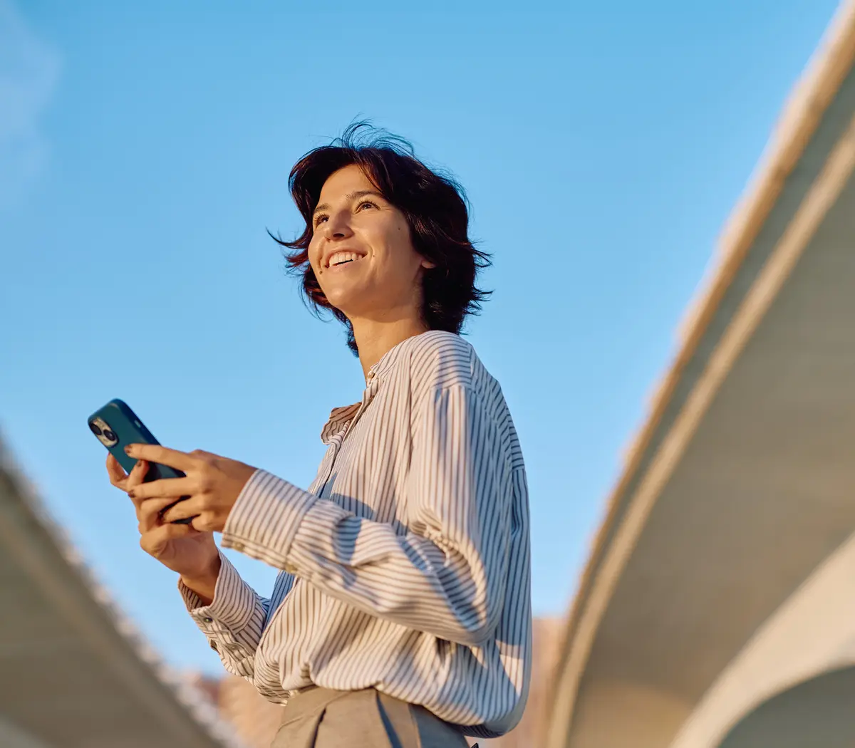 Femme souriante, avec un smartphone en main, sous un ciel bleu.