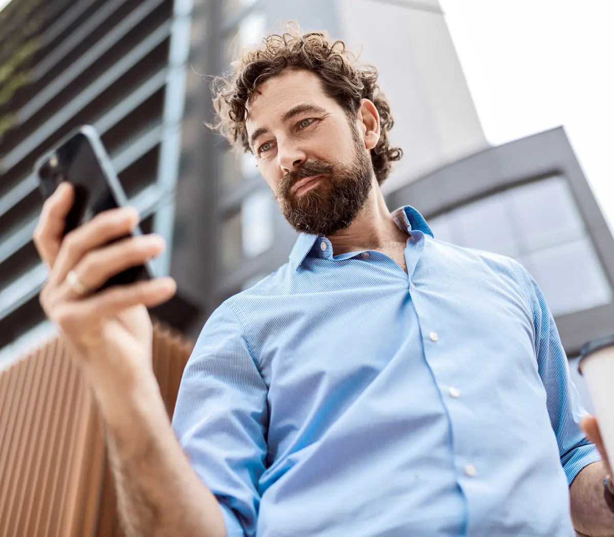 Un homme consulte son téléphone et tient un café devant un bâtiment.