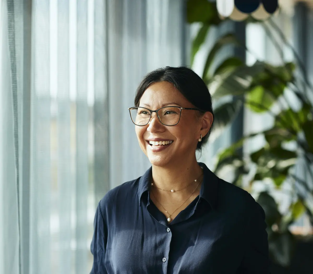 Une femme à lunettes souriante, près de sa fenêtre.