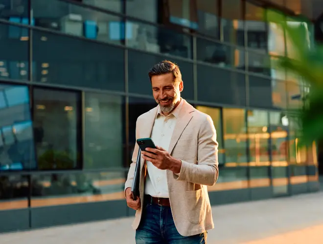 Un homme souriant, avec un téléphone dans les mains, devant un bâtiment moderne.