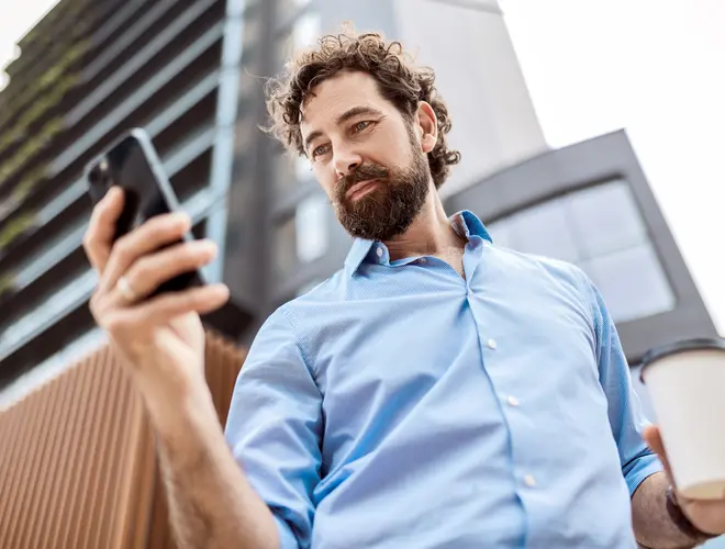 Un homme consulte son téléphone et tient un café devant un bâtiment.