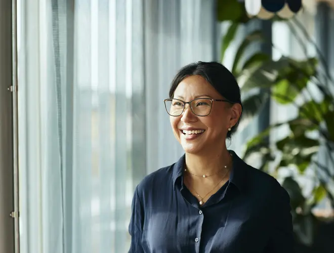 Une femme à lunettes souriante, près de sa fenêtre.