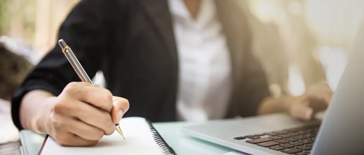 Person sitzt vor Laptop und schreibt im Notizbuch