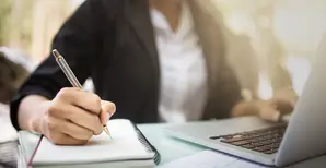 Person sitzt vor Laptop und schreibt im Notizbuch