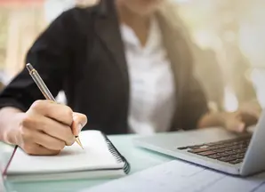 Person sitzt vor Laptop und schreibt im Notizbuch