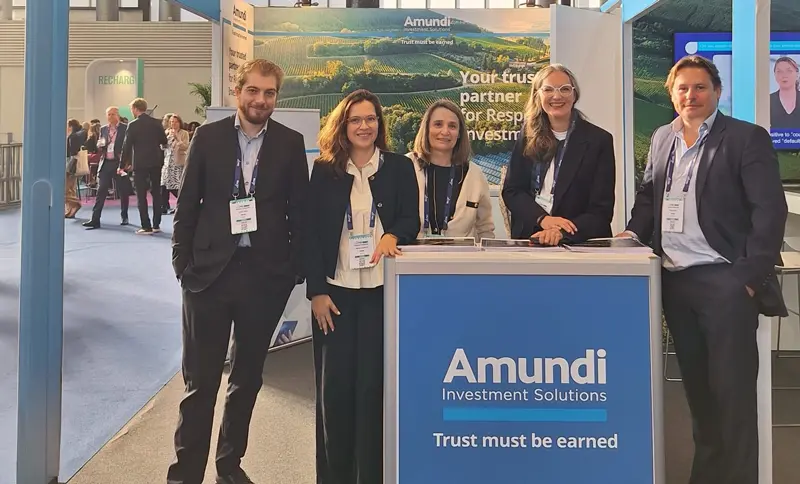 Amundi Investment Solutions booth at a trade fair, with five team members standing behind a counter under a large blue “Trust must be earned” sign. The stand features Amundi branding, exhibition displays and a busy conference hall in the background.