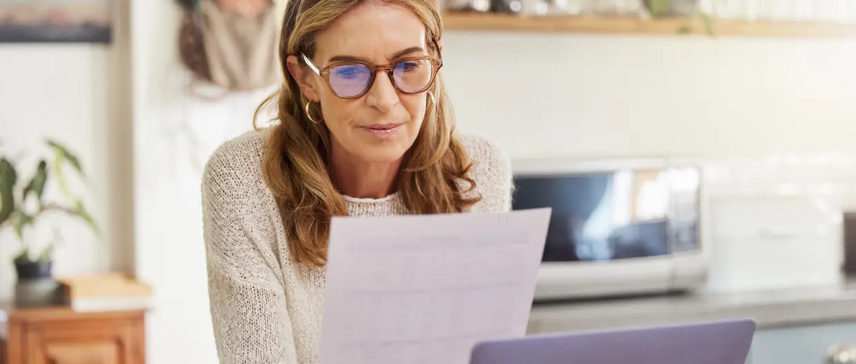 Donna matura con occhiali da vista mentre legge documento cartaceo seduta davanti al computer