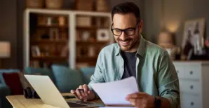 uomo sorridente seduto in salotto mentre trascrive documenti cartacei sul proprio computer