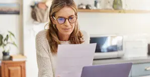 Donna matura con occhiali da vista mentre legge documento cartaceo seduta davanti al computer