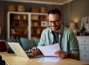 uomo sorridente seduto in salotto mentre trascrive documenti cartacei sul proprio computer