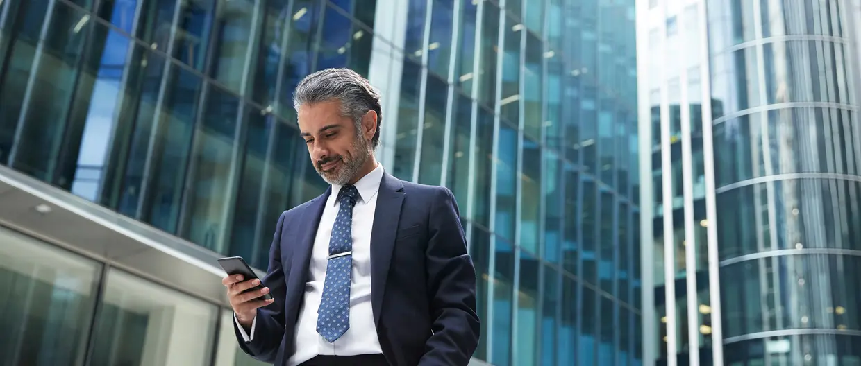 A business man is looking at his phone against tall office buildings.