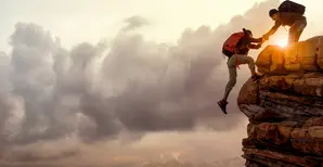 A hiker is being helped by another hiker on a cliff against a sunset.