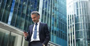 A business man is looking at his phone against tall office buildings.