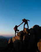 zwei Personen, die bei Sonnenuntergang auf einen Gipfel klettern und sich gegenseitig helfen