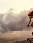 A hiker is being helped by another hiker on a cliff against a sunset.