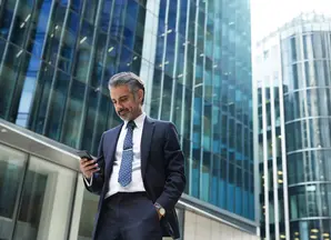 A business man is looking at his phone against tall office buildings.