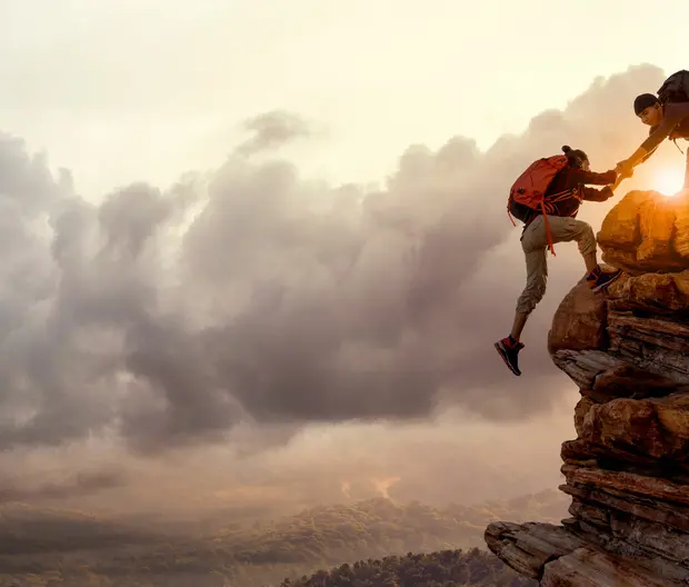 A hiker is being helped by another hiker on a cliff against a sunset.