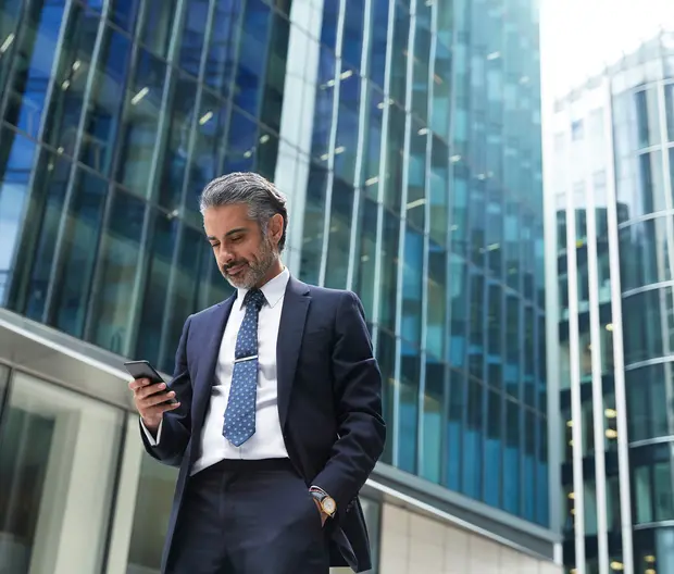 A business man is looking at his phone against tall office buildings.
