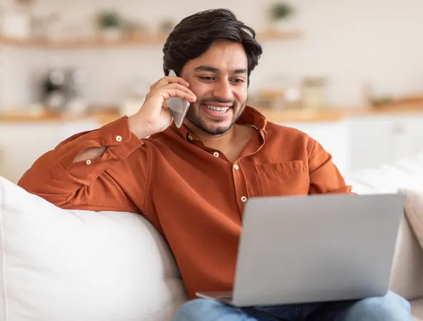 man in his couch on the phone and looking at his laptop 