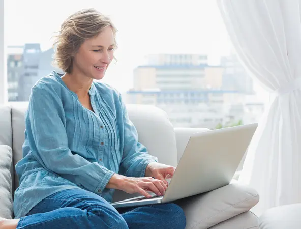 femme blonde assise sur son canapé tapotant sur son ordinateur portable 